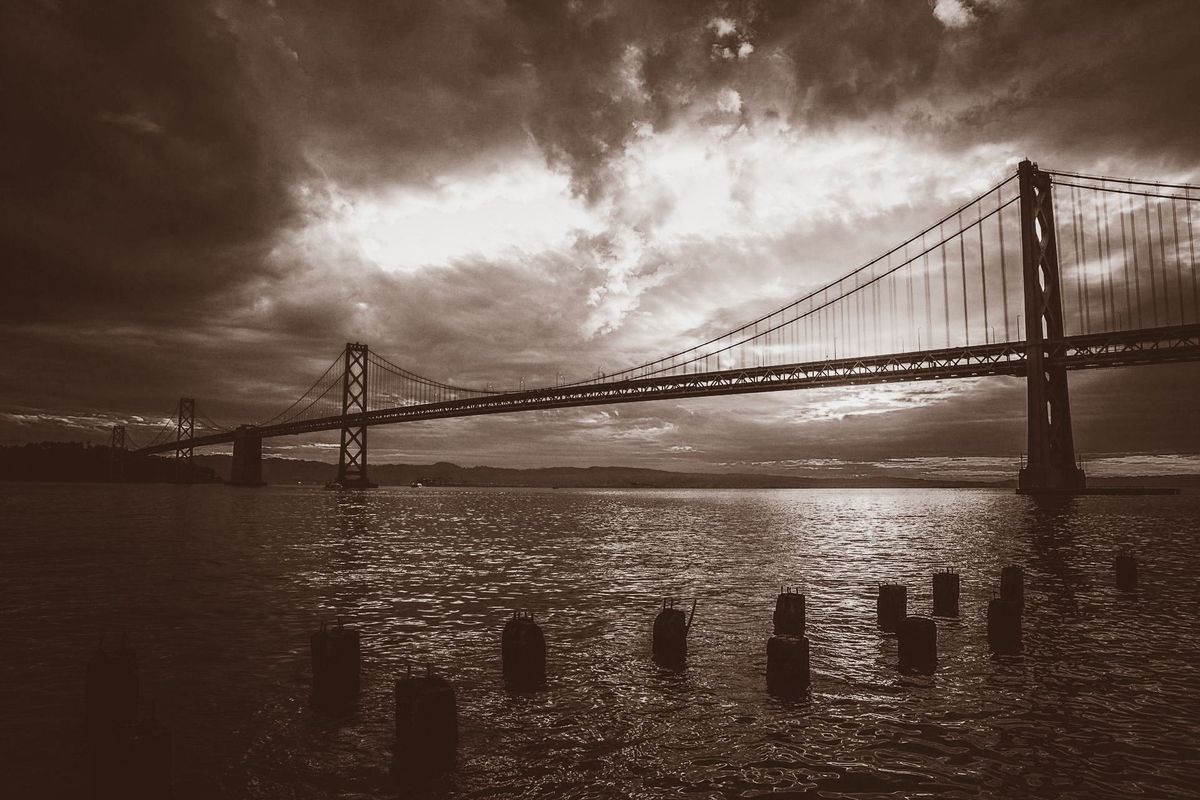 Suspension bridge over water at dusk — dark dramatic clouds, wooden posts in foreground water, sepia / brown tone